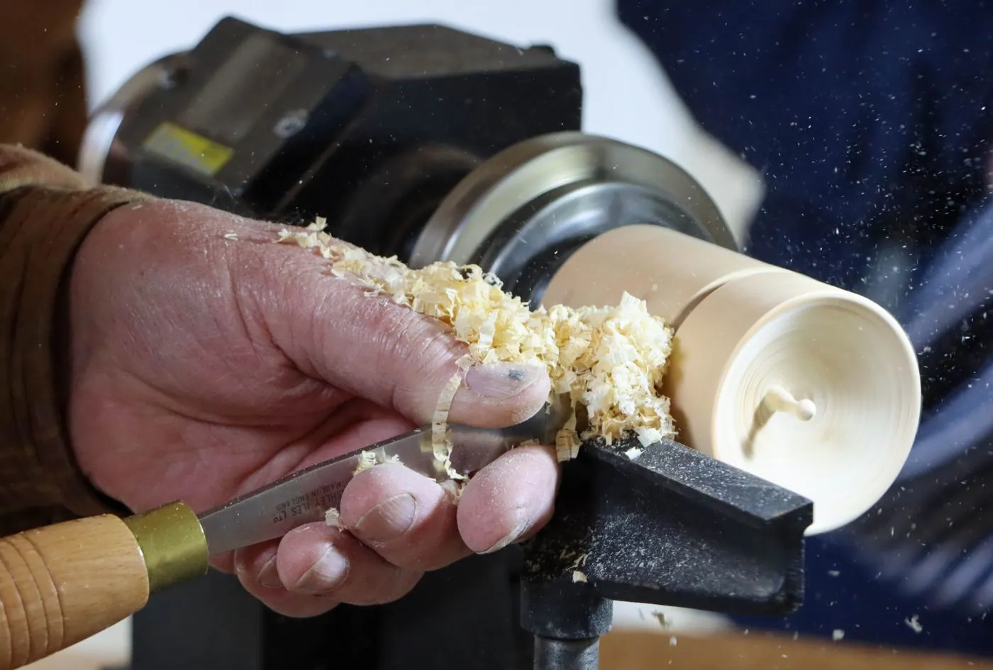 A turner's hand holding a chisel against a pale workpiece on the lathe while shavings fly from the cut
