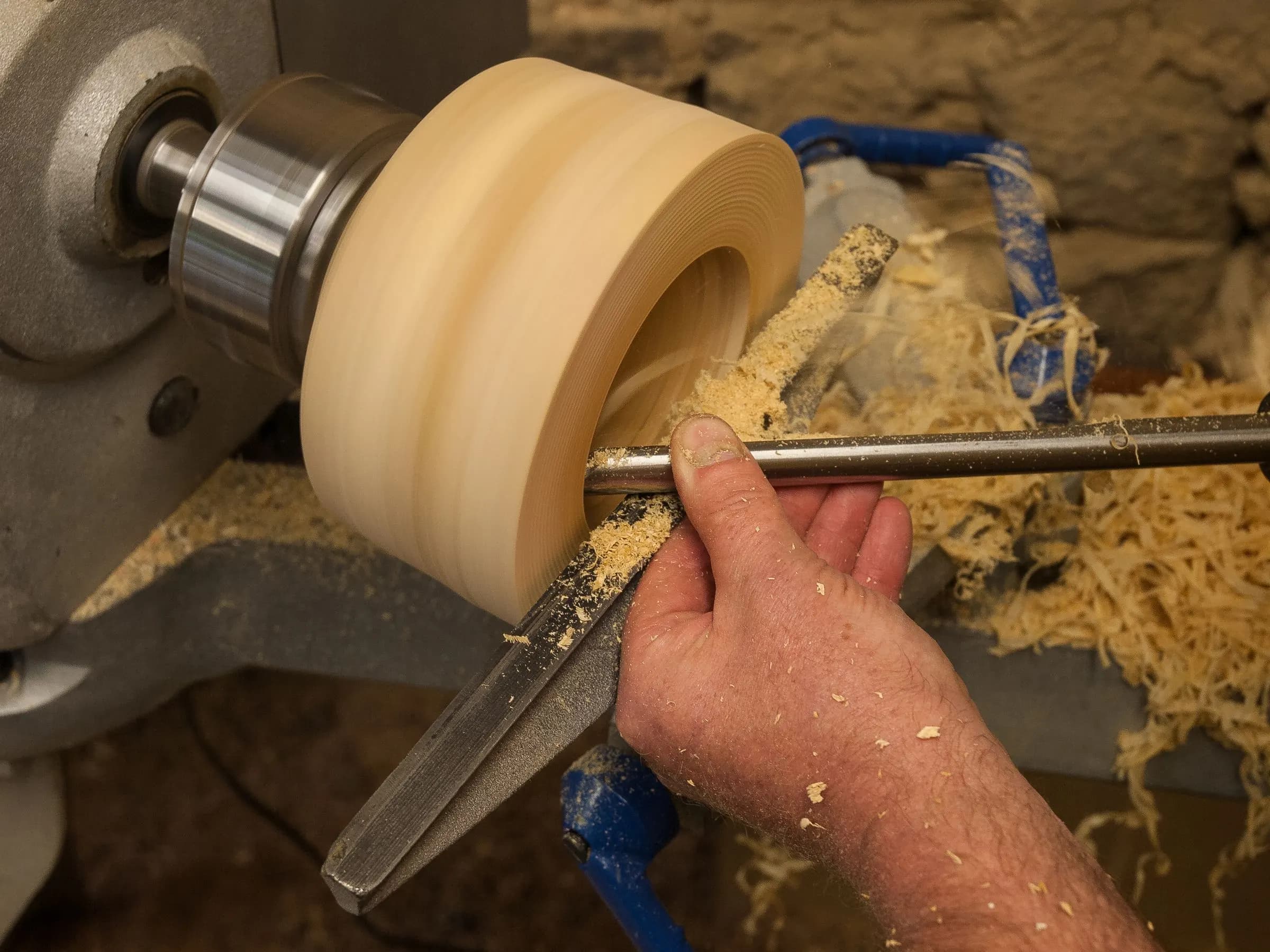 A woodturner's hand holding a tool against a pale bowl blank spinning on a lathe, with curls of shaving thrown off the cut